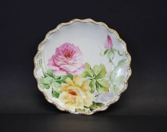 May include: A white, scalloped-edge plate with a hand-painted floral design. The plate features a large pink rose, a yellow rose, and a pink rosebud, with green leaves. The rim is trimmed with gold. A decorative piece for display.