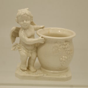May include: A white ceramic figurine of a cherub holding a white ceramic vase with a grapevine design. The vase is decorated with raised grape clusters.