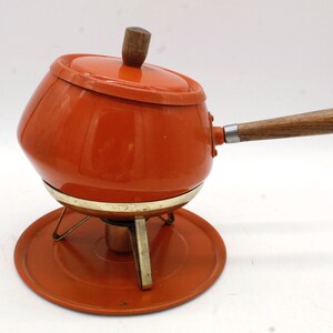 May include: An orange fondue pot with a wooden handle and lid. The pot sits on a metal stand with a matching orange base. The handle and lid knob are made of wood.