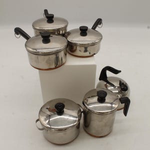 May include: A collection of stainless steel pots with lids and black handles. The pots are of varying sizes and are arranged on a white surface. The lids have black knobs. The pots are designed for cooking.
