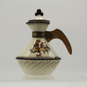 May include: A white ceramic creamer with a rooster design and a wooden handle. The creamer has a woven pattern on the bottom and a gold band around the neck.