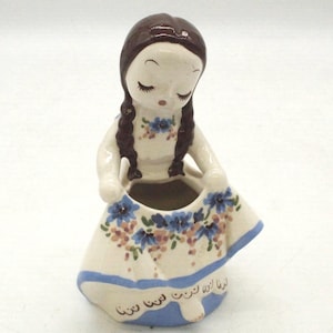 May include: A ceramic figurine of a girl with brown hair in braids, wearing a white dress with blue floral accents. The dress has a blue border with text. The figurine is a decorative item.