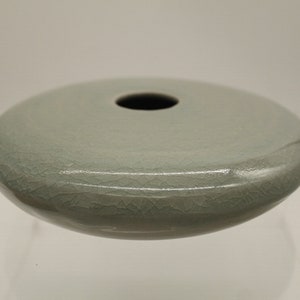 May include: A small, light green ceramic vase with a round, flat base and a small opening at the top. The vase has a crackled glaze.