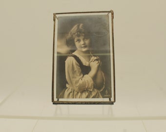 May include: A framed sepia-toned photograph of a young girl with her hands clasped together. The girl is wearing a white dress with a dark bodice and a white collar. The photo is framed in a clear glass frame with a gold metal trim.