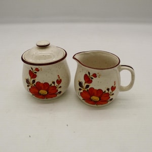 May include: A cream-colored ceramic sugar bowl with a lid and a creamer. Both pieces are decorated with a red floral design and brown trim. The sugar bowl has a round shape, and the creamer has a handle and spout.