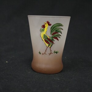 May include: A frosted glass drinking glass with a brown ombre effect. The glass features a hand-painted rooster design with yellow, green, and red colours.