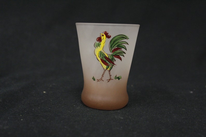 May include: A frosted glass drinking glass with a brown ombre effect. The glass features a hand-painted rooster design with yellow, green, and red colours.