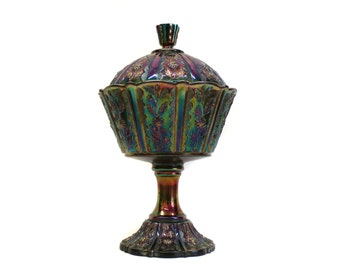 May include: A pedestal candy dish with a lid, crafted from iridescent carnival glass. The piece features a deep purple and green metallic finish with embossed floral patterns and vertical fluting on both the bowl and the circular base.