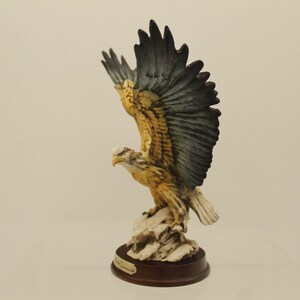 May include: A porcelain figurine of a bald eagle with outstretched wings. The eagle is perched on a rock base with a brown wooden base. The eagle is painted in shades of yellow, brown, and black.