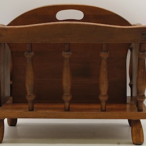 May include: A wooden magazine rack with a curved top and a handle cutout. The rack has a rectangular base and four curved legs. The back features three vertical spindles. The wood has a warm, medium brown tone.