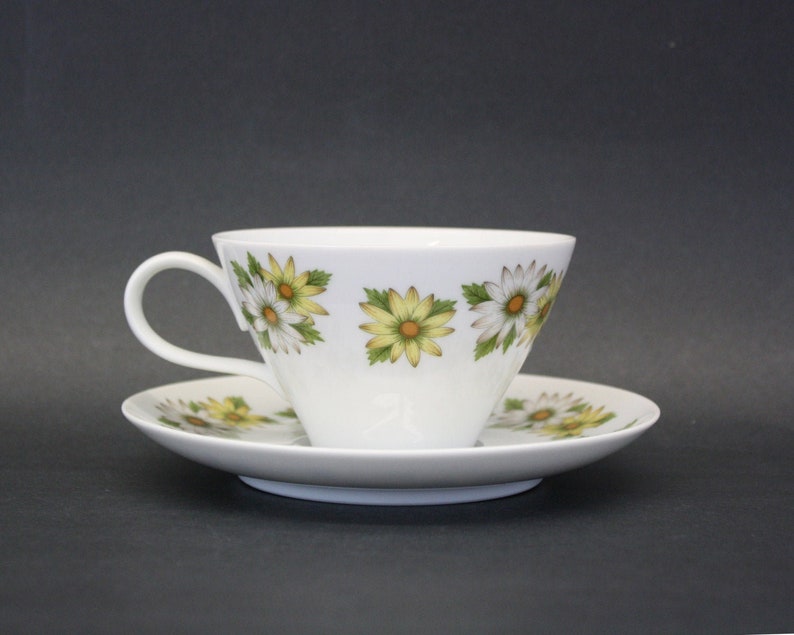 Puede incluir: Juego de taza y platillo blancos con un estampado floral de margaritas en amarillo, blanco y verde. La taza tiene un asa curvada y el platillo es redondo. El juego es probablemente de porcelana.