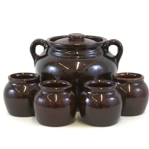 May include: A set of brown ceramic kitchenware. The set includes a large pot with a lid and two handles, and five smaller matching pots. The pots have a glossy finish and are arranged on a white background.