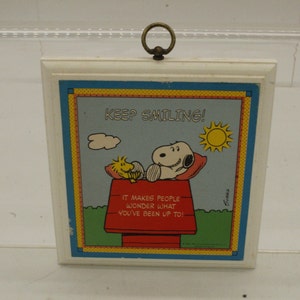 May include: A white wooden plaque with a colorful image of Snoopy and Woodstock. The plaque says "Keep Smiling!" and "It makes people wonder what you've been up to!"