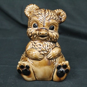 May include: A brown ceramic teddy bear figurine with black eyes and paw prints. The bear is sitting with its paws in front of it.