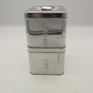 May include: A two-tiered, stainless steel container for coffee and tea. The top section is labeled "COFFEE" vertically, and the bottom section is labeled "TEA". The container has a rectangular shape with a lid.
