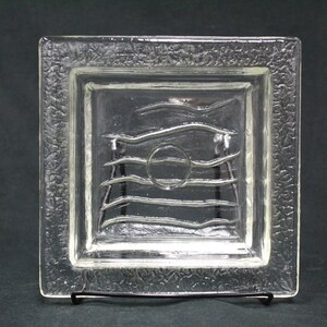 May include: A clear glass square dish with a textured border and a raised design of wavy lines and a circle in the center.