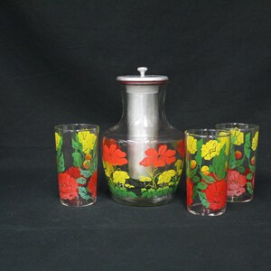May include: A clear glass pitcher with a silver lid and three matching glasses. The pitcher and glasses are decorated with red, yellow, and green floral designs. The set is likely for serving beverages.