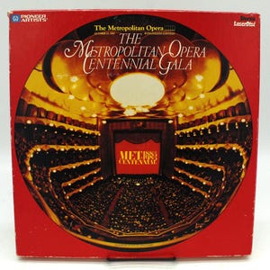 May include: A square LaserDisc cover with a red background featuring a circular image of the Metropolitan Opera house interior. The text reads The Metropolitan Opera Centennial Gala, Pioneer Artists, and Met Centennial in gold and white lettering.