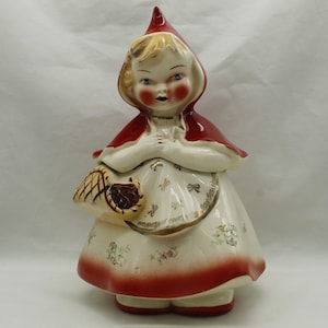 May include: A ceramic cookie jar in the shape of Little Red Riding Hood. The jar features a red hood and dress trim, a white apron with floral and gold accents, and a basket. The jar is a vintage collectible.