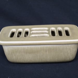 May include: A rectangular ceramic ashtray with a light brown glaze. The ashtray has a series of small holes on the top for extinguishing cigarettes. The bottom of the ashtray has a ribbed design.