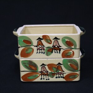 May include: Two square ceramic containers with handles. The containers are stacked on top of each other. Both containers have a white background with a painted design of green leaves, orange circles, and black and pink flowers.