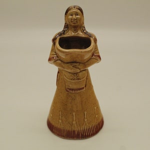 May include: A ceramic figurine of a Native American woman holding a bowl. The figurine is painted in shades of brown and red, with intricate details on the woman's clothing and the bowl.