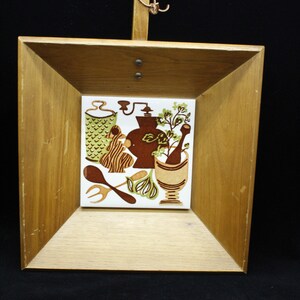 May include: A wooden framed tile with a brown, green, and white design of kitchen utensils and herbs. The tile is hanging from a wooden handle.