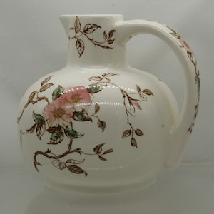 May include: A white ceramic pitcher with a floral design. The pitcher has a round body and a handle. The floral design features pink roses and brown leaves.