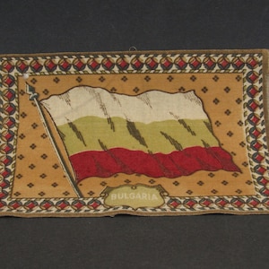 May include: A vintage fabric patch with a depiction of the Bulgarian flag. The flag is white, green, and red with a gold border. The patch has a brown background with a gold and red border. The text "BULGARIA" is printed in gold below the flag.