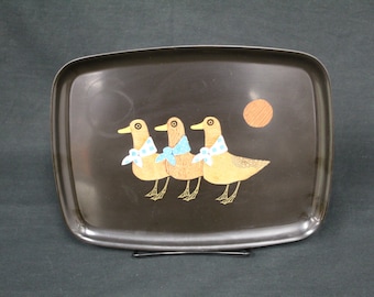 May include: A black metal serving tray with a design of three ducks wearing blue and white scarves. The ducks are brown and yellow and have black eyes. There is a small orange circle in the background.