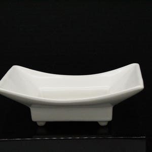 May include: A white, square-shaped ceramic dish with a gently curved rim. The dish is elevated on a rectangular base with three small, rounded feet. The overall design is minimalist and modern, suitable for serving or display.