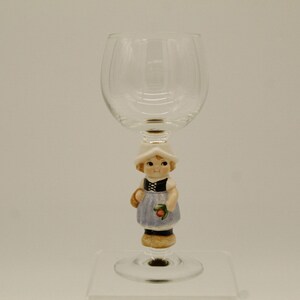 May include: A clear glass wine glass with a ceramic figurine of a girl in a blue dress and white hat attached to the stem.