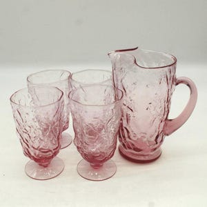 May include: A pink glass pitcher and four matching glasses. The pitcher has a handle and a textured surface. The glasses have a pedestal base and a similar textured design. The set is ideal for serving beverages.
