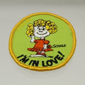 May include: A green and yellow embroidered patch featuring a cartoon girl with blonde hair and a red dress. The text "I'm in love!" is written in black below the girl. The text "©1971 Schulz" is written in white in the upper left corner of the patch.