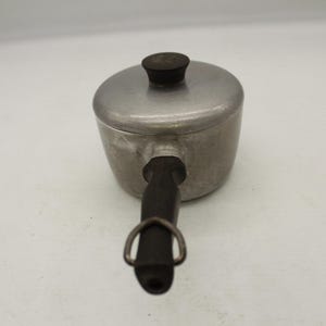 May include: A small, vintage aluminum saucepan with a lid and a dark wooden handle. The handle has a metal ring for hanging. The lid has a dark wooden knob. The pot is likely used for cooking.