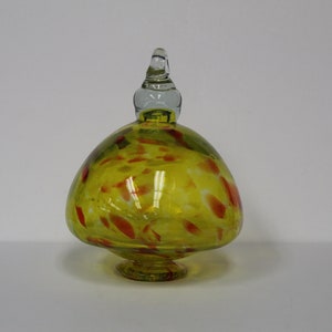 May include: A yellow glass ornament with red and white flecks. The ornament has a clear glass finial on top and a round base.