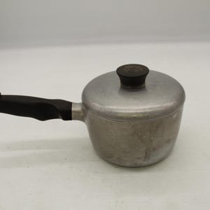 May include: A small, silver-colored saucepan with a lid and a black handle. The handle is attached to the side of the pot, and the lid has a black knob. The pot is made of metal and appears to be vintage.
