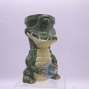 May include: A ceramic alligator-shaped planter in shades of green, yellow, and blue. The planter features a detailed textured design, with the alligator's head forming the top opening. The alligator is in a sitting position, with its tail curled.