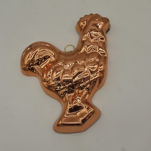 May include: A copper-colored rooster-shaped mold. The mold has a detailed design, with textured feathers and a small loop at the top for hanging. The rooster is a kitchenware item.