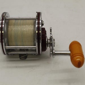 May include: A vintage fishing reel with a silver frame, maroon accents, and a light-colored spool of line. The reel features a bright orange handle and intricate mechanical details, suggesting durability and craftsmanship.