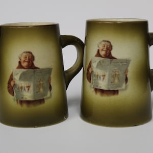 May include: Two green ceramic mugs with a brown glaze. Each mug features a cartoon image of a man in a brown robe reading a newspaper. The newspaper has the word "MAY" printed on it.