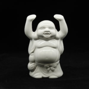 May include: White ceramic figurine of a smiling Buddha with arms raised in a gesture of welcome. The figurine is standing on a black surface.