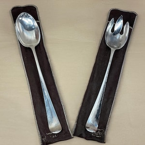 May include: A silver serving spoon and fork set. The spoon has a large, oval bowl and a long handle. The fork has three tines and a long handle. Both are in dark brown protective sleeves.