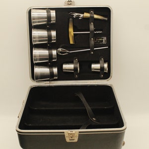 May include: A black travel bar set with a silver metal case. The case is open and contains six silver shot glasses, a silver bottle opener, a silver corkscrew, a silver cocktail stirrer, and a silver ice bucket.