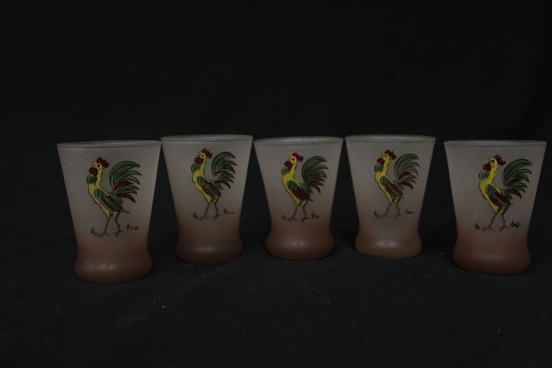 May include: Set of five frosted glass shot glasses with a rooster design. Each glass features a yellow rooster with green and red accents on a brown background.