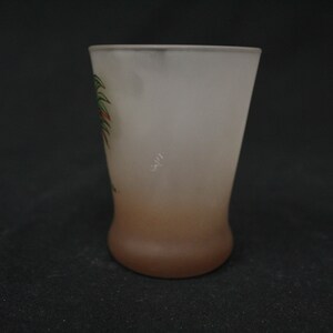 May include: A frosted glass shot glass with a brown ombre effect and a painted design of a palm tree with red and green leaves.
