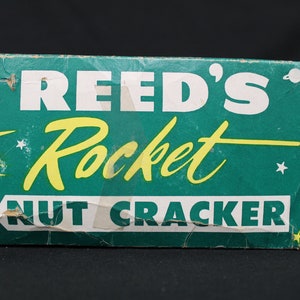May include: Vintage Reed's Rocket Nut Cracker box. The box is teal with white and yellow text. The words "REED'S Rocket NUT CRACKER" are displayed. The box has a retro design with a star and planet graphic.