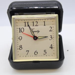 May include: A square analog travel alarm clock with a cream-colored face, black Arabic numerals, and the brand name Equity printed in black. It features black hour and minute hands, a red second hand, and is housed in a black hinged case.