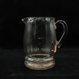May include: A clear glass pitcher with a pink tint and a rounded body. The pitcher has a handle and a wide base.