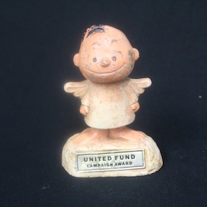 May include: A small, beige ceramic figurine of a smiling angel with wings. The angel is standing on a base with the words "UNITED FUND CAMPAIGN AWARD" inscribed on a silver plaque.